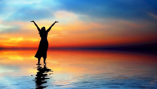 a woman raises her arms in worship on a shoreline facing a sunset