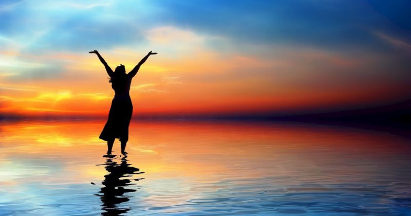 a woman raises her arms in worship on a shoreline facing a sunset